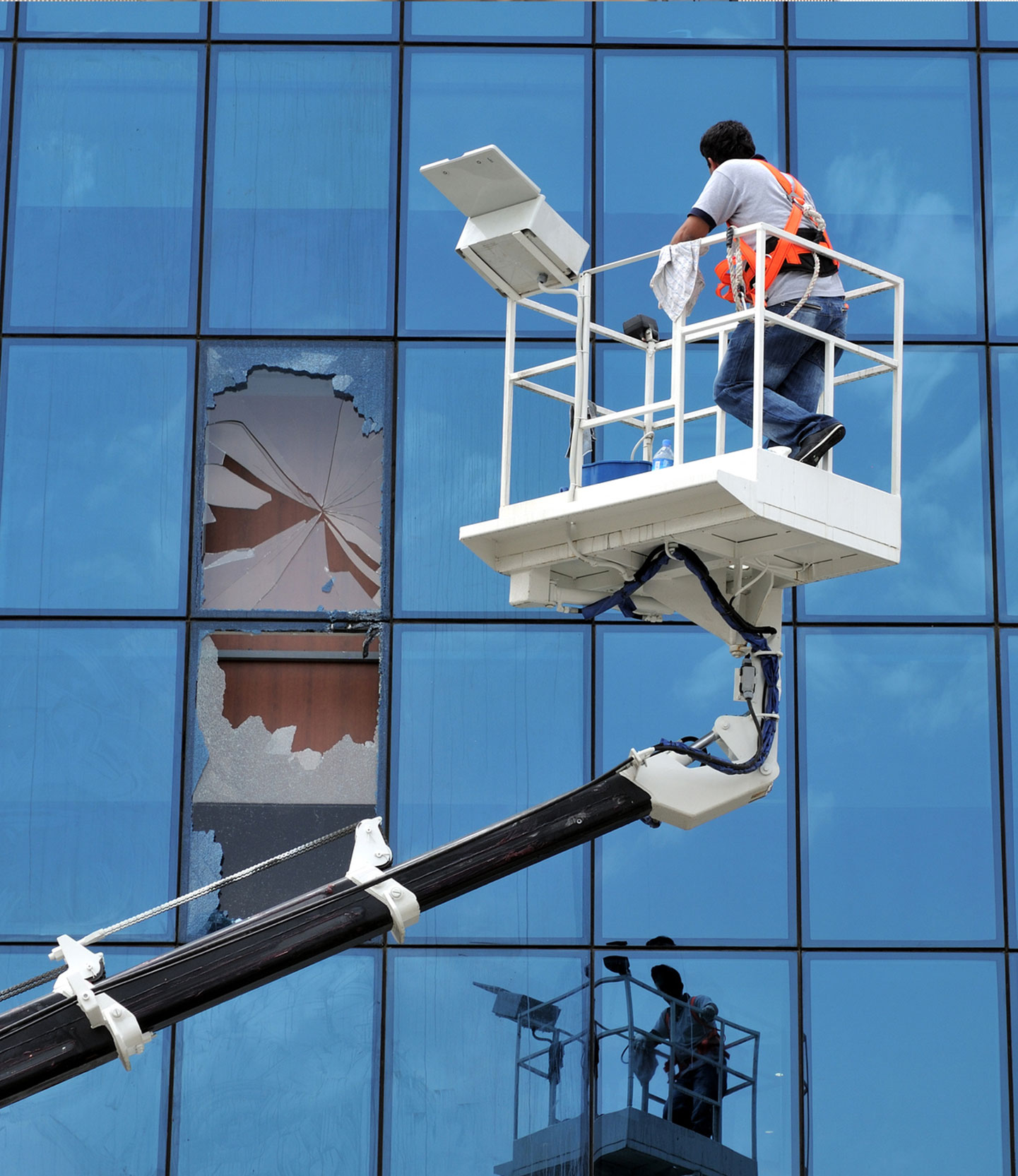 A man on a lift repairing a commercial building that received damage from a storm. The window is shattered and the repair man is fixing it.