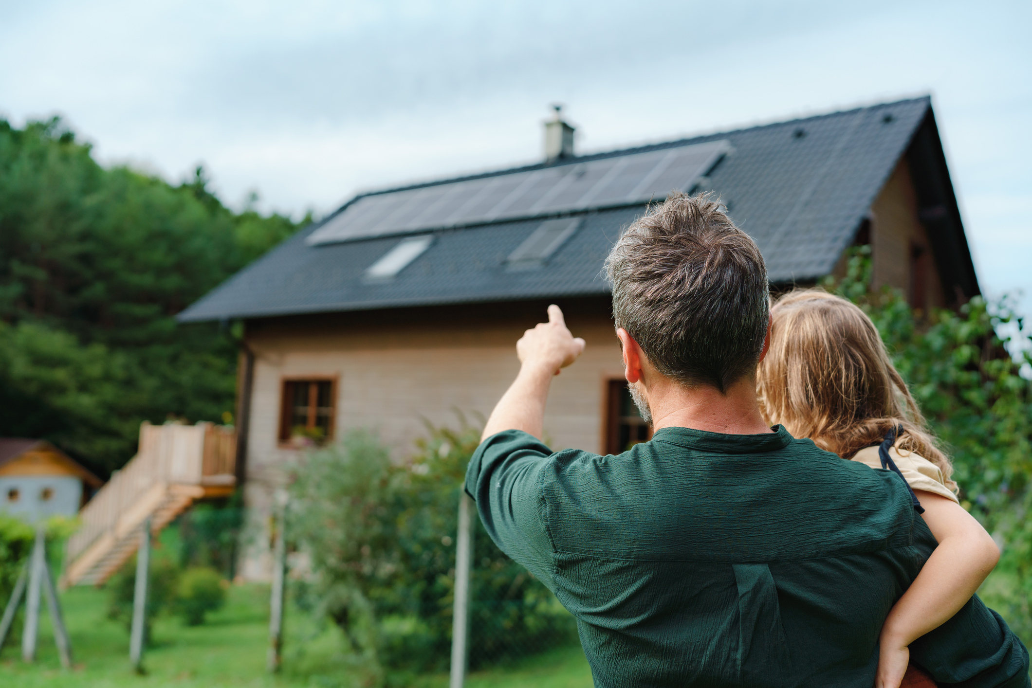 Rear view of dad holding her little girl in arms and showing at their house with solar panels.Alternative energy, saving resources and sustainable lifestyle concept. Their house was just repaired after property loss damage in Grand Rapids.